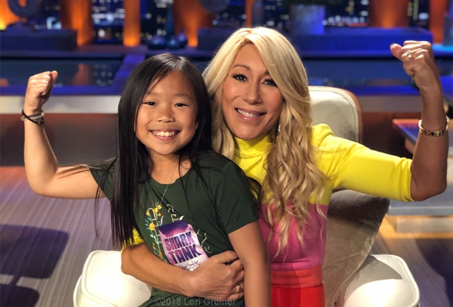 Lori on the set of Shark Tank with a young girl in her lap in a strong arm pose